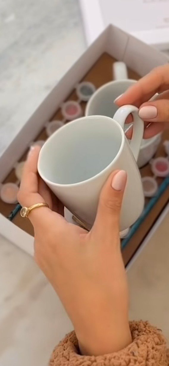 Hands holding a plain white ceramic mug over a tray of paint pots, ready to be painted.